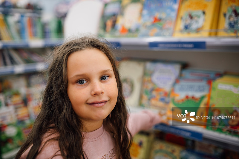 图为一个小女孩在超市里寻找摆满儿童书籍的书架时看着相机图片素材