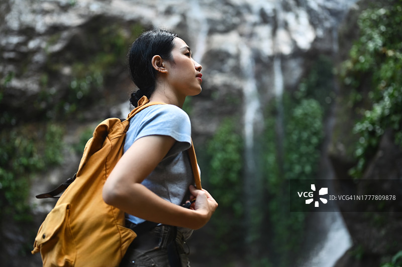 年轻女游客背着背包站在热带瀑布前欣赏自然风光的肖像图片素材