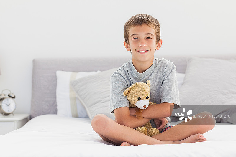 微笑的小男孩坐在床上，抱着他的泰迪熊图片素材