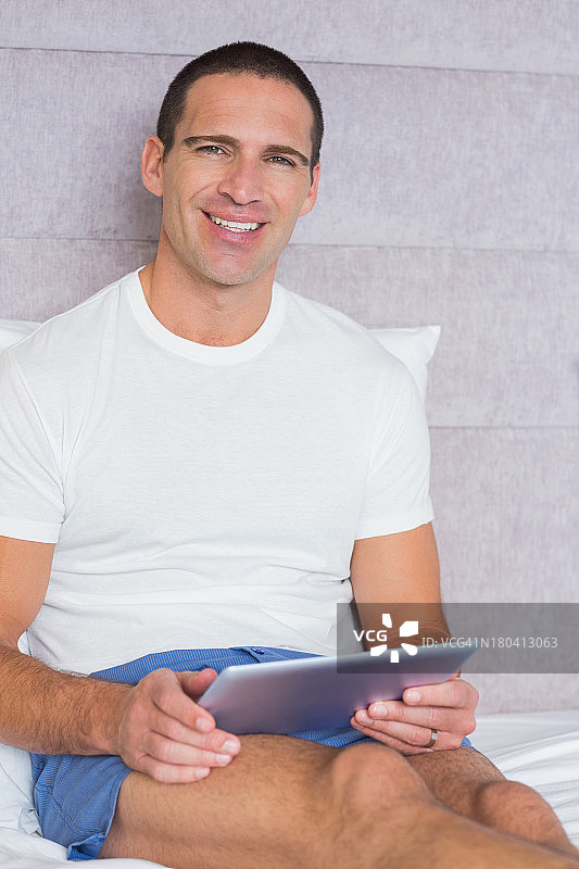 快乐的男人坐在床上用他的平板电脑图片素材