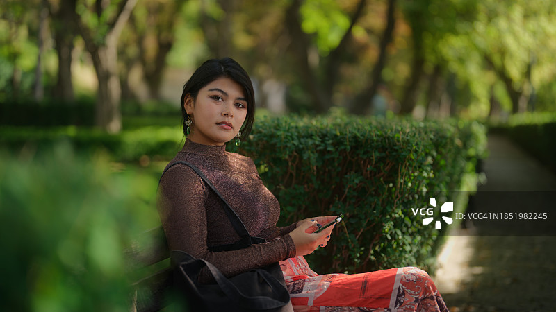 年轻美丽的多民族女性坐在公园长椅上，一边使用手机一边休息图片素材