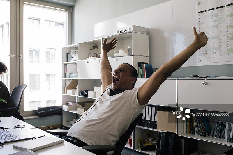 快乐的男性企业家坐在办公室里，举起手臂尖叫图片素材