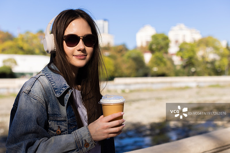 一个女孩走在河堤上，听着音乐，喝着咖啡，看着她的智能手机，春天图片素材