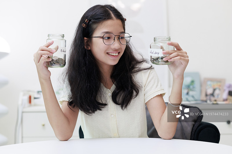 一个年轻的亚洲女孩把硬币放进一个罐子里，象征着她为教育和未来的假期存钱的承诺。图片素材