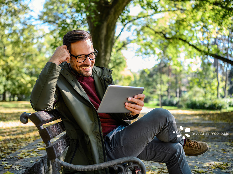 一个面带微笑的男人坐在公园的长椅上，用着一台数码平板电脑图片素材