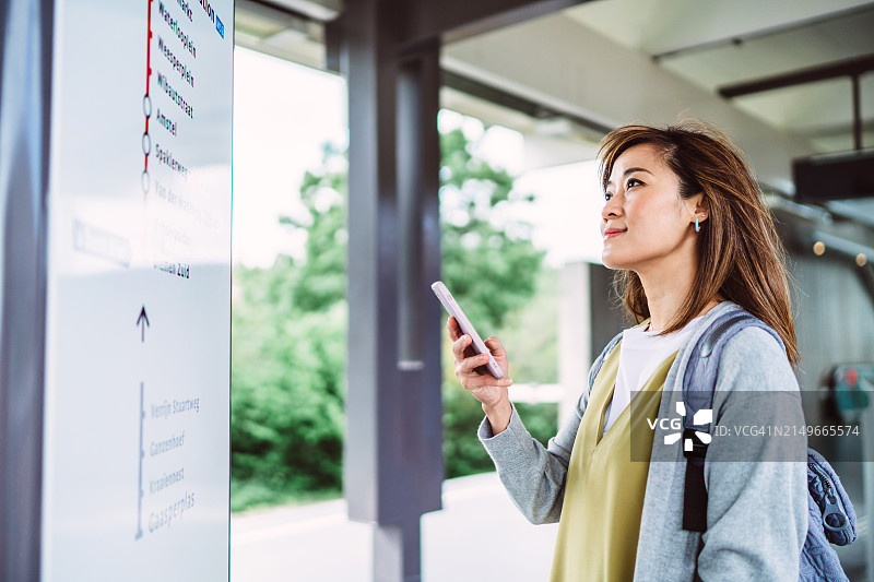 规划旅程:年轻的亚洲女子在探索一个新城市的同时，拿着智能手机在火车站台上查看路线图图片素材