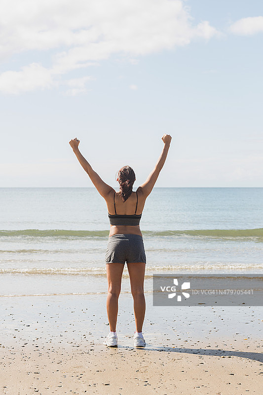 后视图苗条的年轻女子举起她的手臂站在海滩上图片素材
