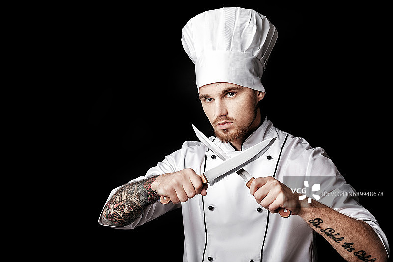 年轻的大胡子厨师，身穿白色制服，手持两把刀，背景是黑色图片素材