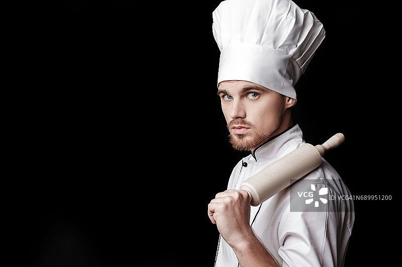 年轻的大胡子厨师，身穿白色制服，手持擀面杖，背景是黑色图片素材