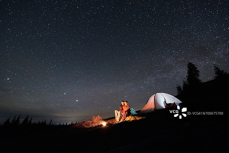 晚上露营。在美丽的夜空下，浪漫的游客夫妇在明亮的帐篷旁的篝火旁休息。天体摄影术图片素材