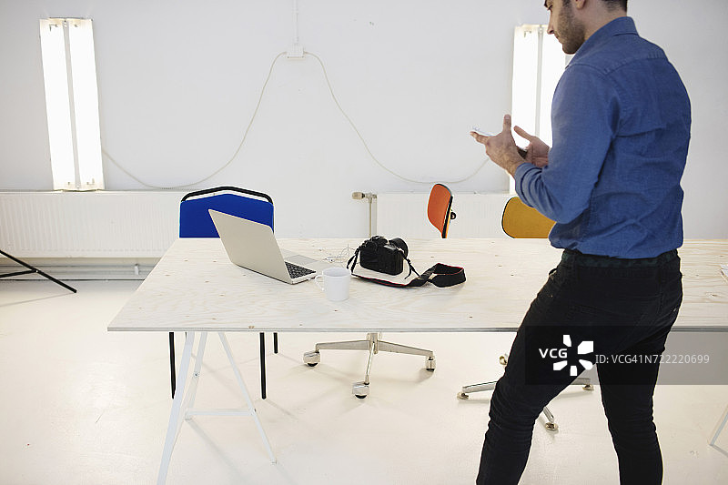 后视图的男性博主使用智能手机，而站在创造性的办公室办公桌图片素材