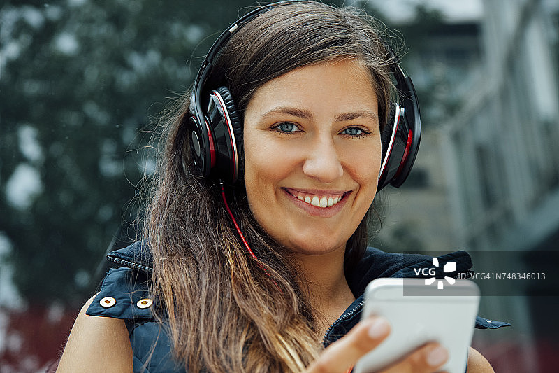 一个微笑的年轻女子的肖像戴着耳机和手机在城市里图片素材