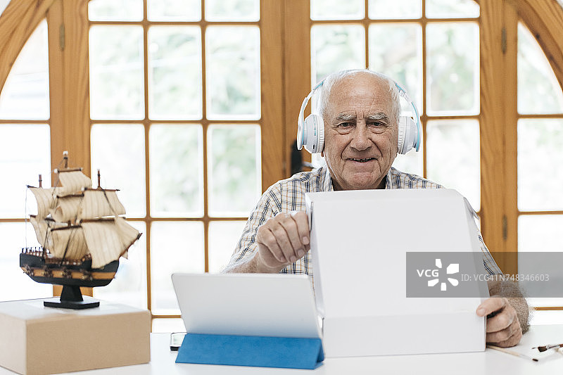 一个戴着耳机的自信的老人的肖像，桌上放着平板电脑，模型船和包装图片素材