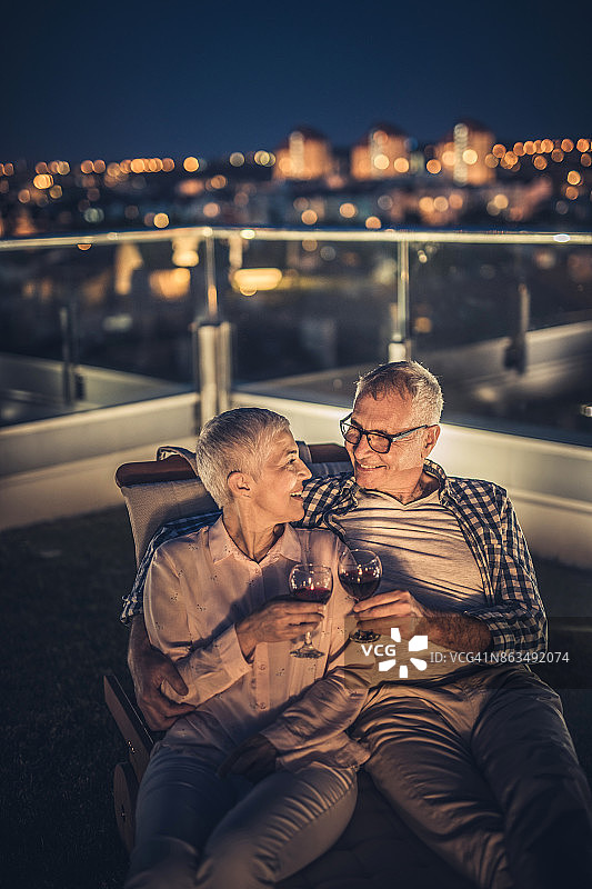 一对相爱的老年夫妇晚上在顶层公寓的露台上用葡萄酒敬酒。图片素材