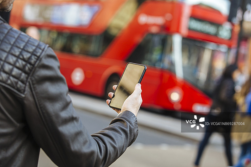 在街道上使用手机，背景是红色的双层巴士，特写图片素材