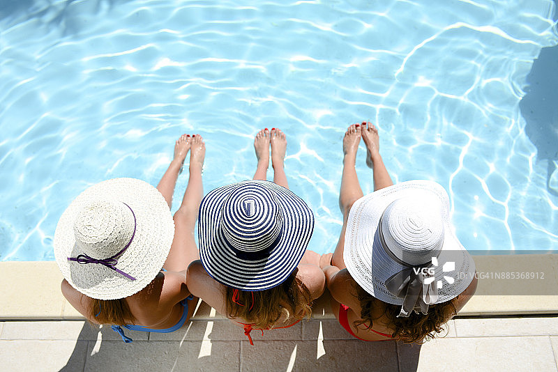 三个漂亮的年轻女子穿着泳衣和太阳帽坐在泳池边度假游泳池在暑假。图片素材