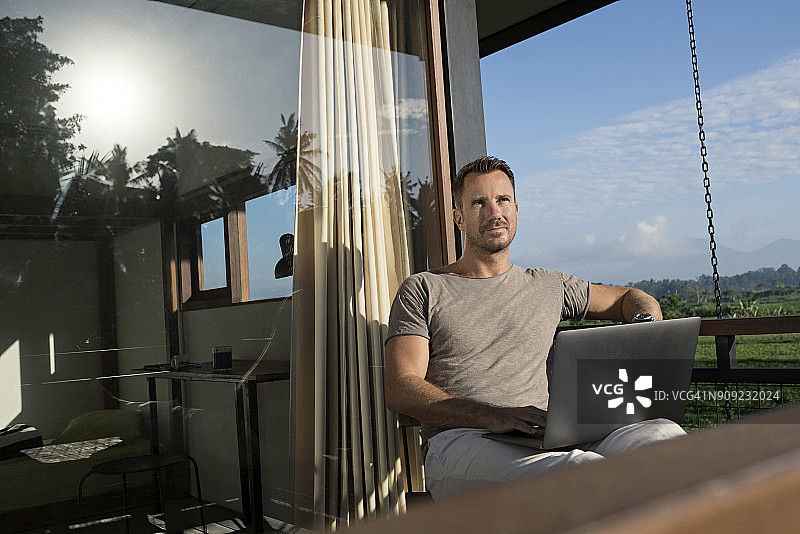 男人坐在现代住宅的阳台上看风景，拿着笔记本电脑图片素材