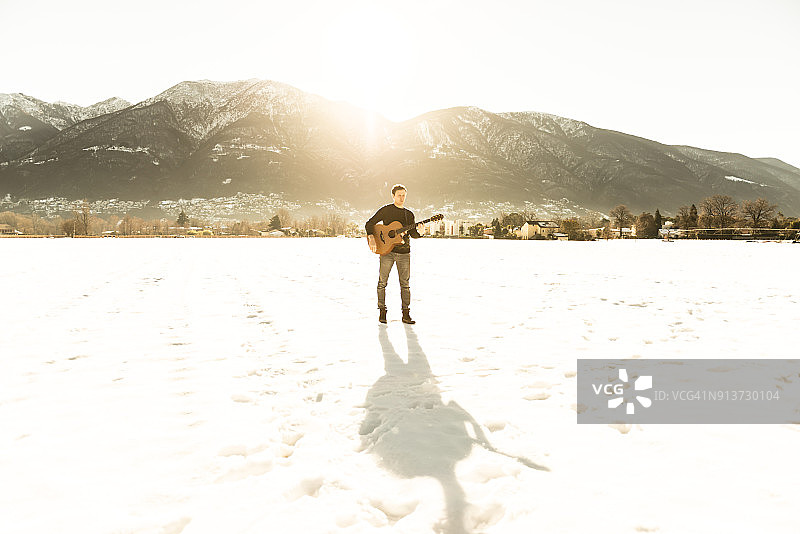 音乐家站在雪地上，带着他的吉他和阴影和阳光越过山图片素材