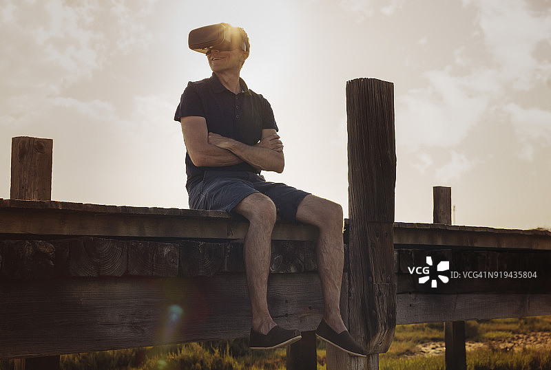 一个戴着VR眼镜的成熟男子坐在木板路上欣赏夕阳下的美景图片素材