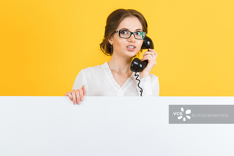 自信美丽兴奋的年轻商业女人的肖像与电话显示空白广告牌在黄色的背景图片素材