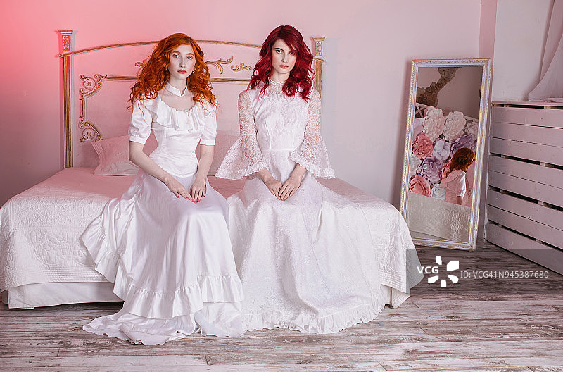 两个红头发的漂亮女孩穿着维多利亚时代的白色婚纱。女性风格。脆弱的女孩。瘦腰。两个女人坐在卧室的一张白色的床上图片素材