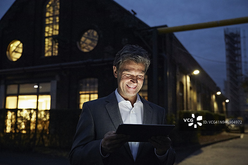 微笑的商人晚上在现代建筑外使用平板电脑图片素材