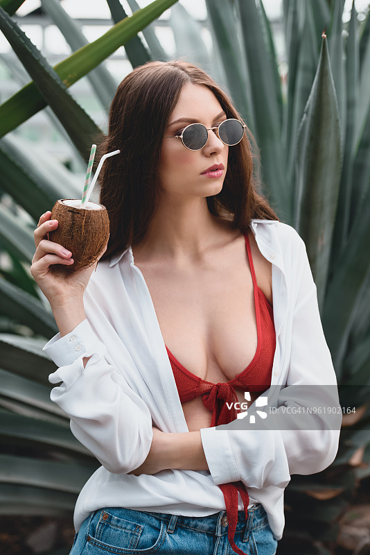 迷人的女孩戴着太阳镜摆姿势与椰子鸡尾酒在热带花园图片素材
