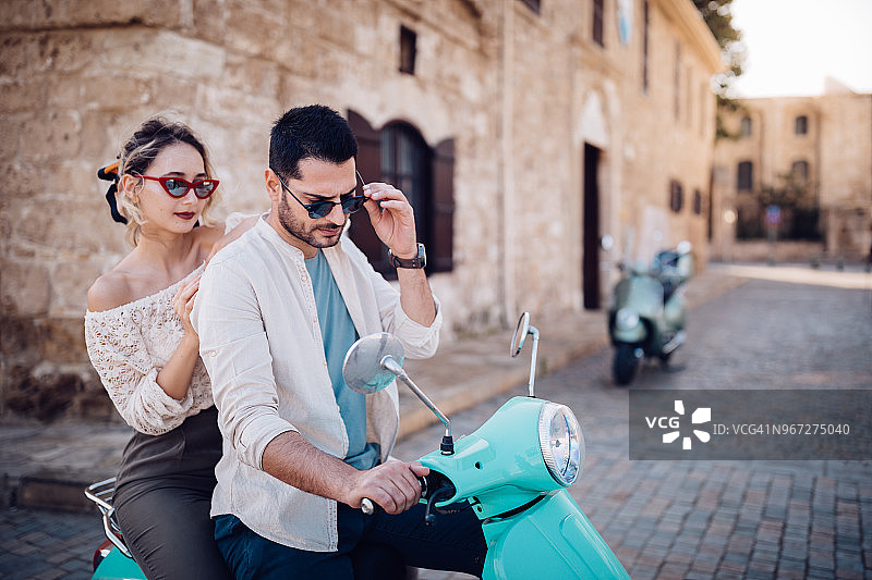 年轻时髦的夫妇骑着滑板车在古老的意大利小镇街道上图片素材