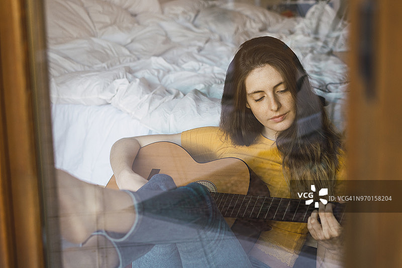 一个放松的年轻女子坐在窗玻璃后面弹吉他的肖像图片素材