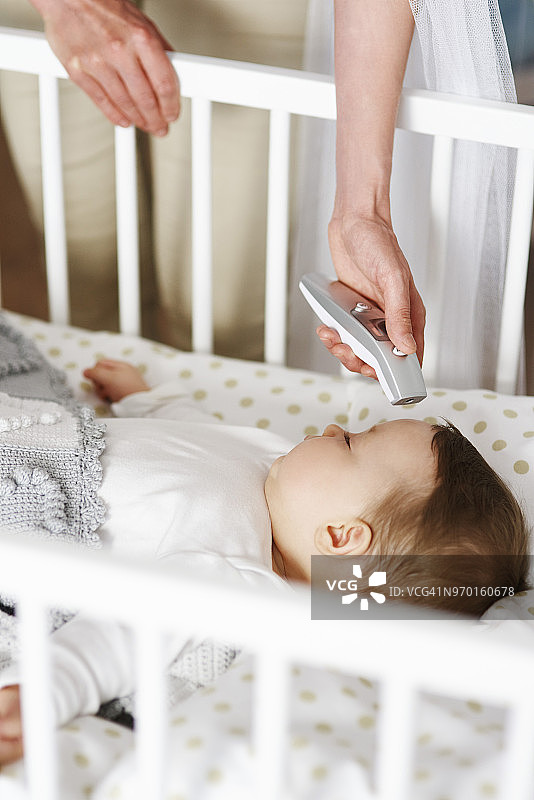 母亲用数字温度计检查婴儿在婴儿床的温度图片素材