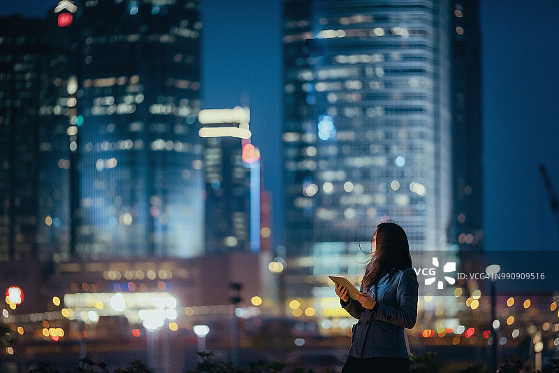 年轻的女商人在夜晚使用数码平板电脑，自信地回顾着灯火通明的城市天际线图片素材