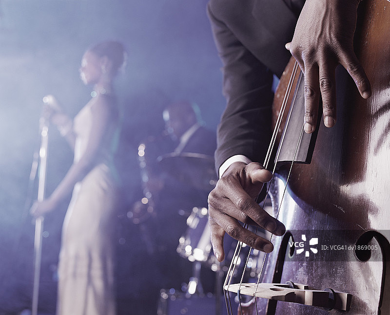 一个男人在一个夜总会的舞台上弹拨一个低音Double Bass和一个女歌手和萨克斯手站在背景的特写图片素材