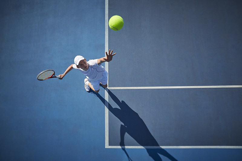 头顶上的年轻男子网球运动员在阳光明媚的蓝色网球场上打网球，发球图片素材