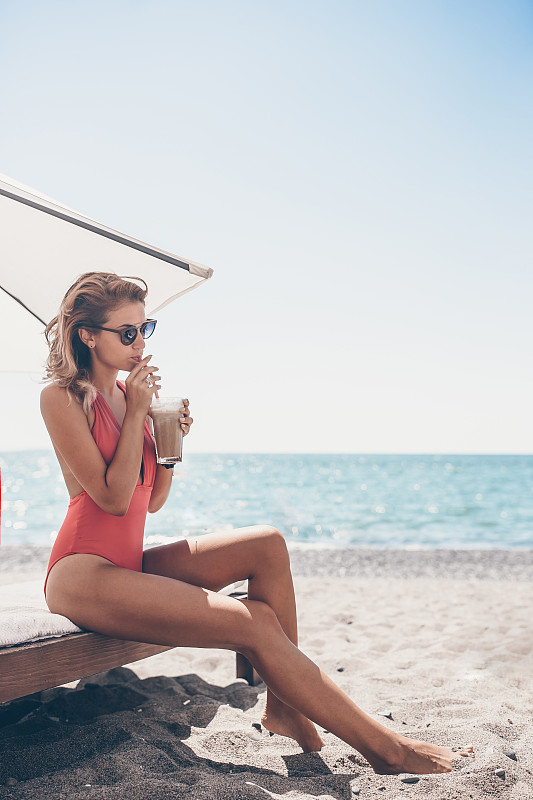 年轻女子拿着鸡尾酒杯坐在白色沙滩上的日光浴床上图片素材