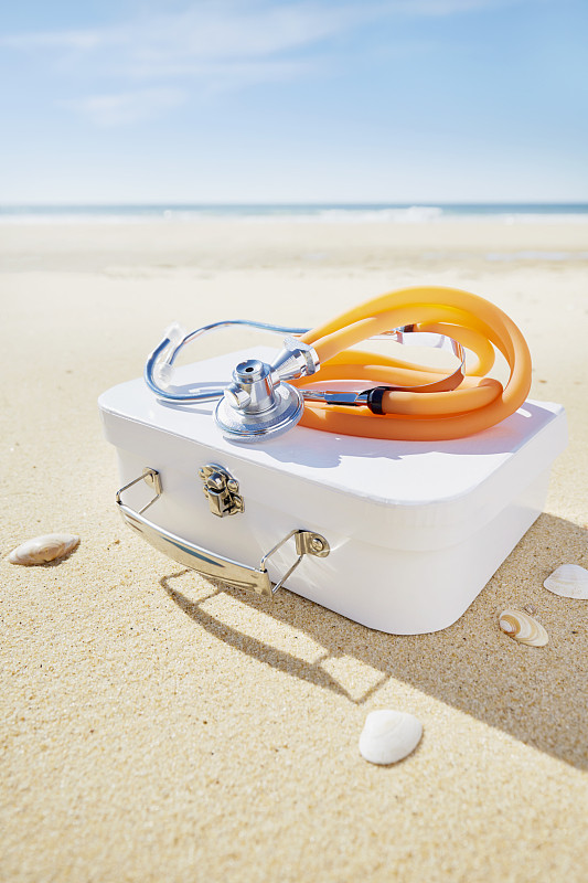 海边沙滩上的听诊器和手提箱，旅行药房/急救箱符号图片下载