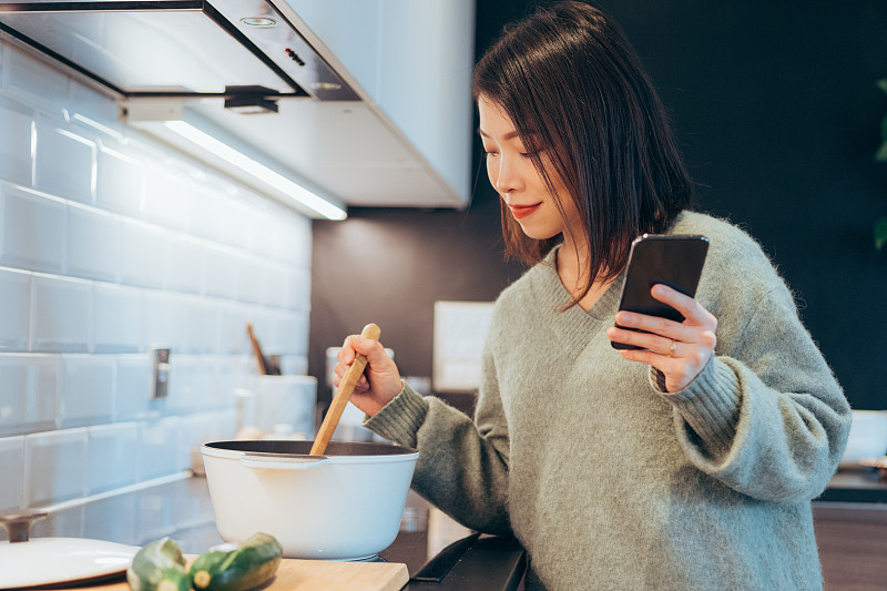 年轻快乐的女人拿着智能手机在家厨房做饭图片素材