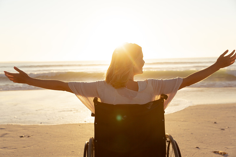 妇女在轮椅上的后视图与她的双臂在海滩上张开图片素材