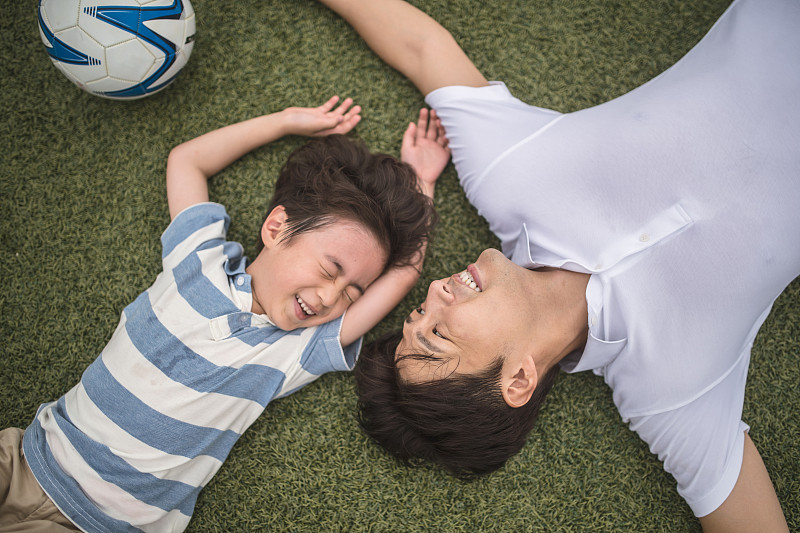 父亲和小儿子伸开双臂躺在足球旁边的草地上图片下载