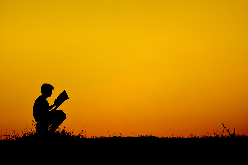 天空日落时，一个正在看书的男孩的剪影图片下载