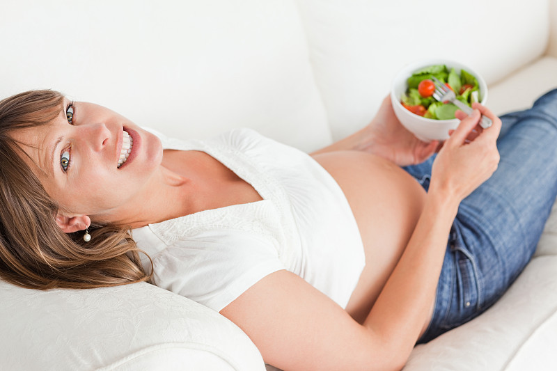 可爱的孕妇躺在公寓的沙发上吃沙拉图片下载