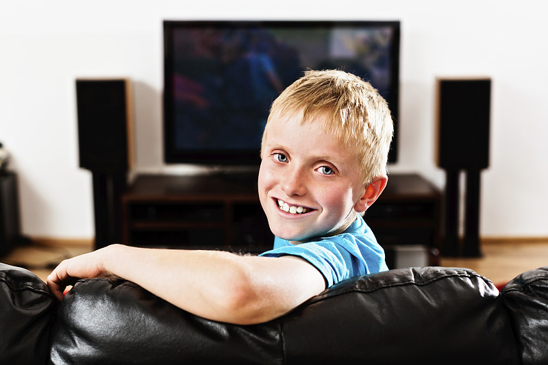 坐在黑色皮沙发上的小男孩从电视上转过身来，微笑着图片下载
