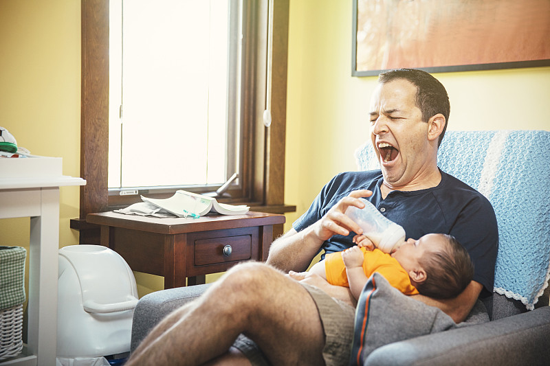打呵欠的白人父亲在客厅给男婴喂奶图片下载