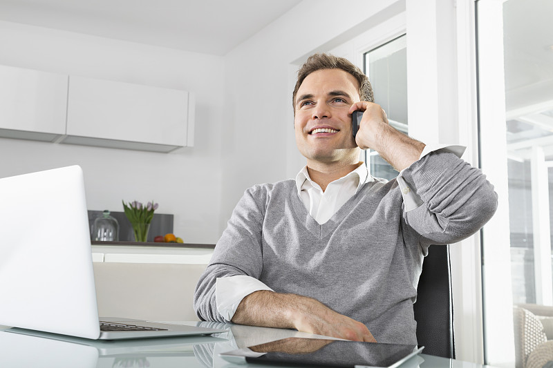 微笑的男人坐在厨房里，拿着笔记本电脑和数字平板电脑，用智能手机打电话图片下载