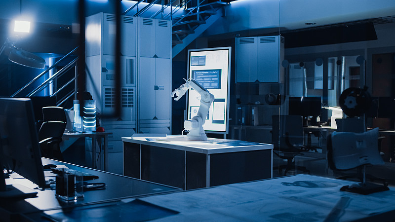 工业机器人设计研究实验室:机器人手臂原型站在桌面照明。在黑暗的背景中各种高科技设备，计算机，数字白板和蓝图图片素材