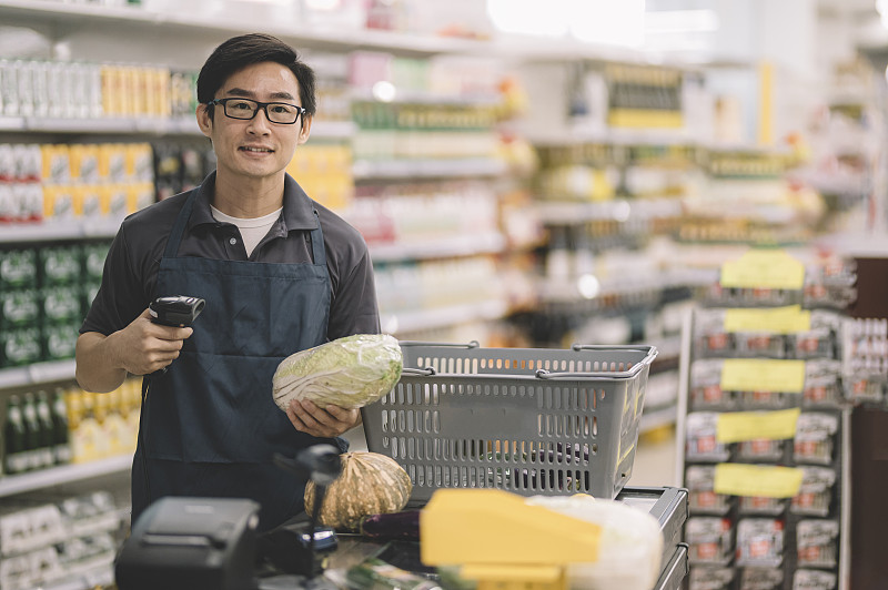 一位亚洲华人销售收银员拿着蔬菜、卷心菜和条形码阅读器站在收银台的收银台前，微笑着看着摄像头图片素材