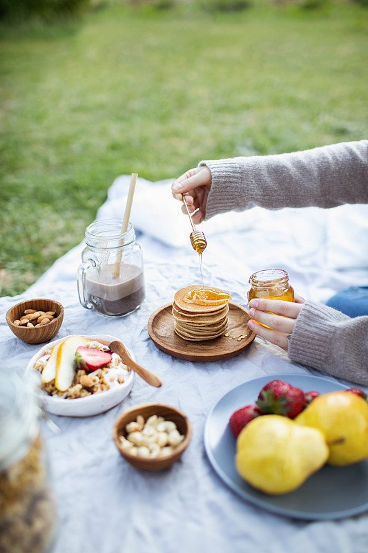 夏季健康素食野餐在公园。
女人用手把有机蜂蜜倒在美味的煎饼上。
水果，可可，蜂蜜煎饼和有机格兰诺拉麦片在亚麻桌布上装饰枕头和柳条篮子。图片下载