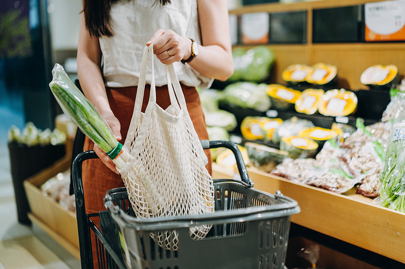 裁剪的镜头年轻的亚洲妇女在超市购买新鲜的有机食品杂货，并把各种水果和蔬菜放入棉花网生态袋在购物车。“零浪费”的概念图片下载