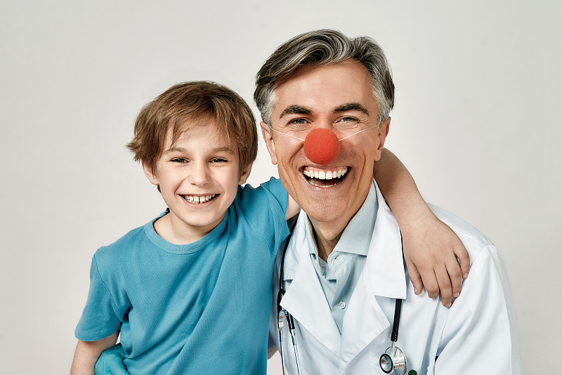 可爱兴奋的小男孩与快乐的长着小丑鼻子的男性儿科医生拥抱的照片。他们看着相机微笑着。与孩子一起工作的医生摄影图片下载