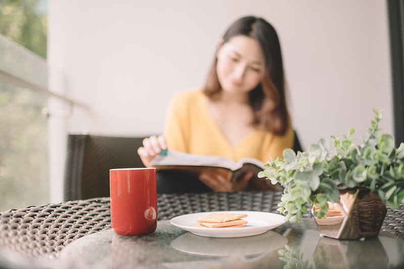 一位亚洲华裔美女中午在她的阳台上看书，放松，享受她的数字排毒时间，喝着咖啡，吃着饼干当点心图片下载