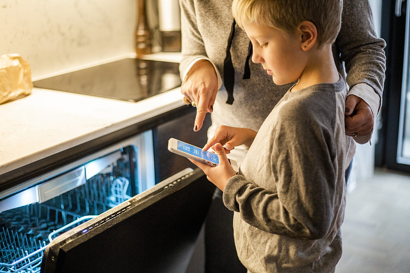 在智能家居的厨房里，男孩通过洗碗机使用手机应用程序，向母亲学习图片下载